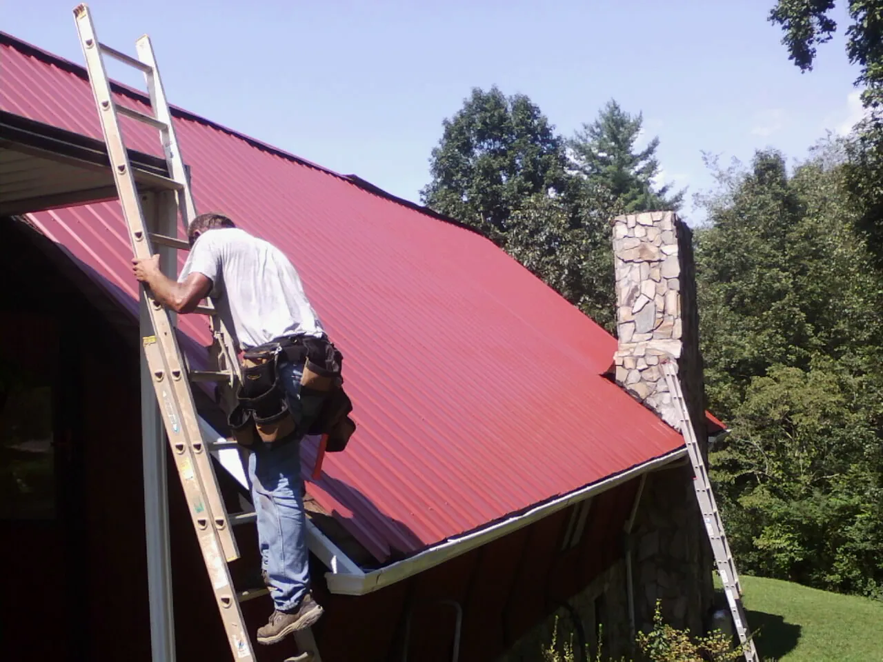 Roofer on a ladder beside a red metal roof and stone chimney — Soffit & Fascia Repair by Roofing Brothers in Boylston